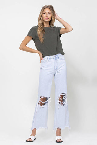 A woman with long, wavy, blonde hair wearing a dark green t-shirt, Aspen Distressed Vintage Ankle Flare Jeans by VERVET by Flying Monkey in light blue ripped denim, and white slide sandals poses with one hand resting on her head and the other hanging by her side, standing against a plain white background.