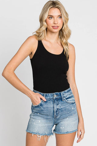 A woman with wavy blonde hair wearing a black sleeveless tank top and Petra153's Stratler High Rise Comfort Short, featuring distressed denim with a frayed hem, stands against a plain white background. Her left hand is in her pocket, and she is looking slightly off to the side.