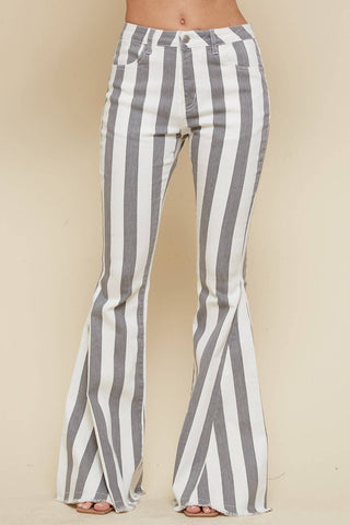 A person is shown wearing WISTERIA LANE's Storm Charcoal Striped Flare Bellbottom Jeans. The pose emphasizes the pants' design, featuring vertical black and white stripes, with a close-up view from the waist to the feet. The background is a neutral beige color.