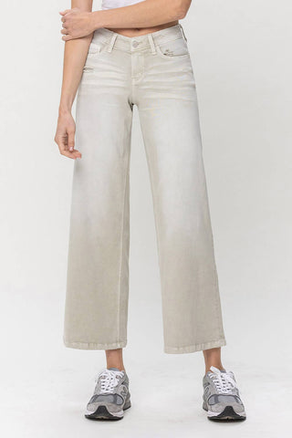 The individual is modeling the Sagelyn Low Rise Baggy Wide Jeans by VERVET by Flying Monkey in light gray, featuring a front button and zipper closure. They are paired with a gray long-sleeve top with rolled-up sleeves and gray sneakers, set against a plain white background.