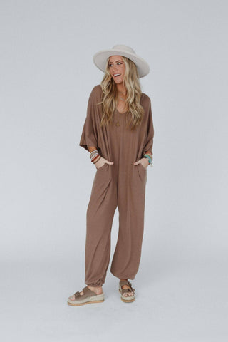 A smiling woman with long blonde hair, dressed in the Lovely As Ever Harem Jumpsuit from Three Bird Nest made from comfortable knit fabric and a wide-brimmed white hat. She is wearing sandals and bracelets on her wrists. Her hands are in her pockets, and she is standing against a plain light grey background.