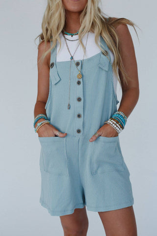 A person with long blonde hair stands against a plain background, wearing the Wild Hearts Button Romper by Three Bird Nest over a white tank top. The light blue striped romper, featuring adjustable straps, is accessorized with multiple beaded bracelets and necklaces. Their hands rest casually in the pockets of the textured romper.