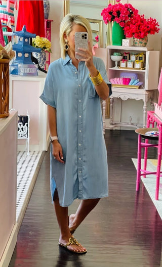 In the enchanting Western Edge Boutique, decorated with vibrant colors and pink flowers, a person takes a mirror selfie wearing an Arrow Denim Button Down Midi Dress. Their look is complete with gold hoop earrings and sandals, showcasing their versatile style amidst shelves filled with delightful items.