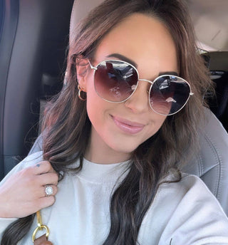 A woman with long wavy hair, slightly smiling, sits in a car wearing DAX Eyewear's Georgie Gold Frame Sunglasses with UV protection. She's in a light sweater and holds a brown handbag.