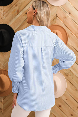 A person with shoulder-length, blonde hair is standing with their back to the camera. They are wearing the Taylor Smocked Cuffed Striped Boyfriend Shirt with Pocket from Aapparella, featuring a light blue and white striped design, a turn-down collar, and long smocked sleeves. The shirt is paired with white pants. Several hats in various colors are hanging on a wooden panel wall in the background.