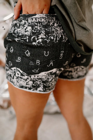 Close-up photograph of a person wearing 2 Fly's SHE GON' COUNTRY black and white patterned tombstone mesh shorts adorned with various illustrations and letters. Their hand rests on the side of their hip, paired with a grey top. The background is blurred and indistinct, evoking the casual vibe of summer events.
