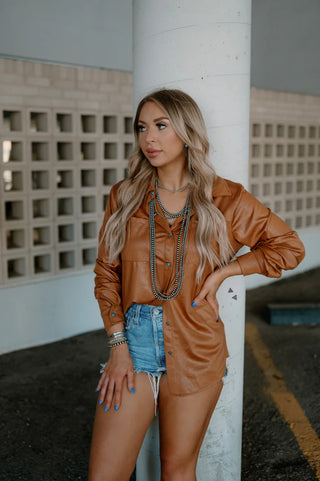 A woman with long blonde hair and blue nail polish stands against a white pillar. She is dressed in the Leather Lux Saddle Brown Long Sleeve Top by Western Edge Boutique, featuring turquoise pearl snaps, layered necklaces, bracelets, and denim shorts. The background showcases a wall with a grid pattern.