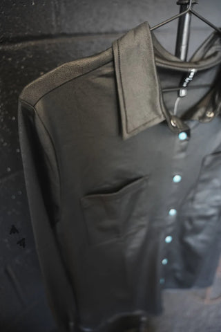 Close-up of a black LEATHER LUX * MIDNIGHT button-down long-sleeve shirt by 2 Fly on a hanger, featuring a single breast pocket and a row of TURQ PEARL SNAPS. The shirt is displayed against a dark textured wall in a clothing store.