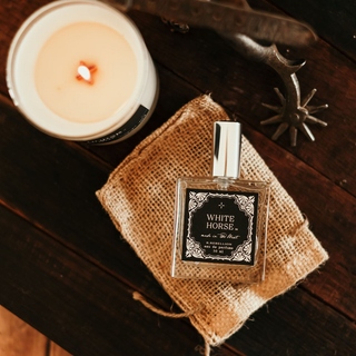 A bottle of "White Horse Perfume 50 ml" by R. Rebellion rests on a burlap drawstring bag on a wooden surface, exuding rugged beauty. Next to it is a lit candle and vintage metal spurs. The label of the perfume bottle showcases an ornate, decorative design.
