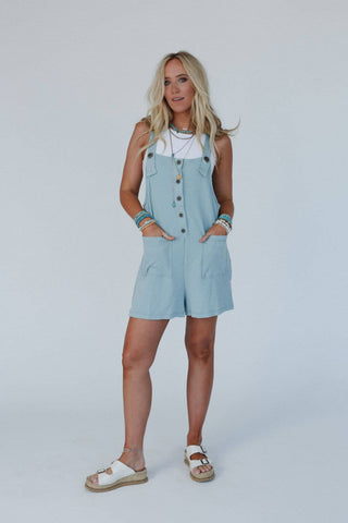 A woman with long blonde hair stands against a plain background wearing the Wild Hearts Button Romper by Three Bird Nest, styled in light blue with a textured pattern, layered over a white tank top. She accessorizes with multiple bracelets and necklaces, completing her look with white sandals. Her hands rest in the romper's pockets, and she is looking forward.