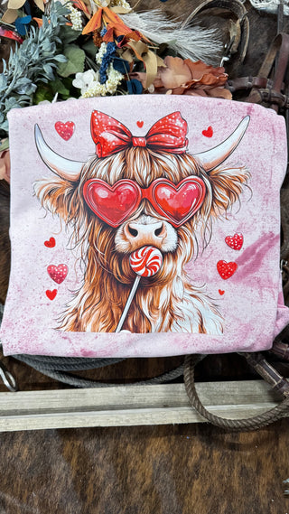 The "Highland Type of Love" design by Western Edge Boutique features a cartoon cow wearing heart-shaped sunglasses, a red polka dot bow, and holding a lollipop among red hearts, set against a pink backdrop with flowers and ropes.