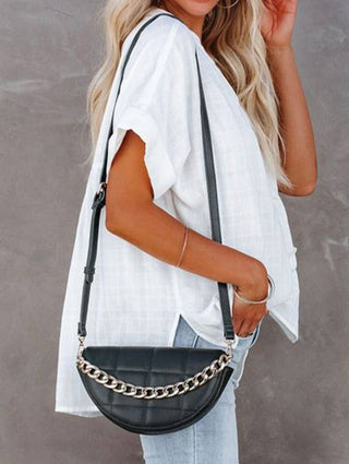 A woman with wavy blonde hair wears the Pretty Bash Aria Loose V Neck Lightweight Summer Women's Top in white, paired with light blue jeans. She carries a black half-moon shaped crossbody bag featuring a silver chain accent. Her hand is casually placed in her jean pocket as she stands against a neutral gray background.