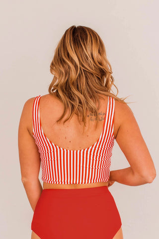 A woman with wavy blonde hair is seen from the back, highlighting the Betsy Front Knot Swimsuit Tankini in a red and white stripe design by Western Edge Boutique. The tankini provides a perfect fit with high-waisted red bottoms and features a tattoo adorning her upper back, adding flair to the sassy ensemble. The background is plain and light-colored.