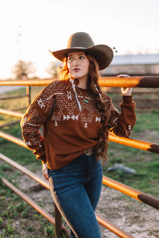 A woman stands by a fence wearing the 2FLY Saddle Ranch Sweater, along with jeans and a wide-brimmed hat. Her long hair flows freely as she gazes into the distance, while the setting sun casts a warm glow over the scene.