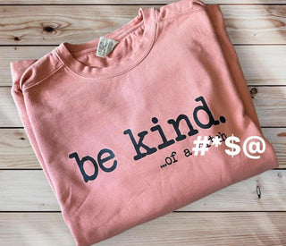 A folded coral Be Kind Sweater from Western Edge Boutique with the words "be kind" printed in large black letters. Below that, smaller text reads "...of a #*$@." The sweater is displayed on a wooden surface featuring a light-colored wood grain pattern.