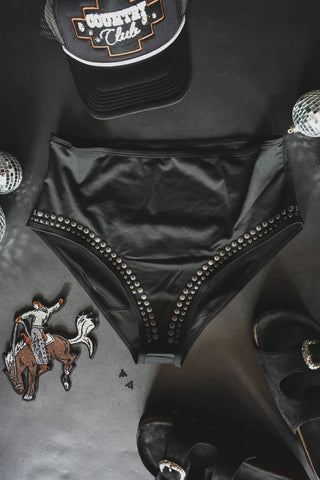 A pair of CABO COWBOY BOTTOMS by 2 Fly, featuring black high-waisted design with silver studs, is placed on a dark surface. Surrounding it are a black cap with "Country Club" text, small disco balls, a rodeo-themed patch, and black high-heeled sandals.