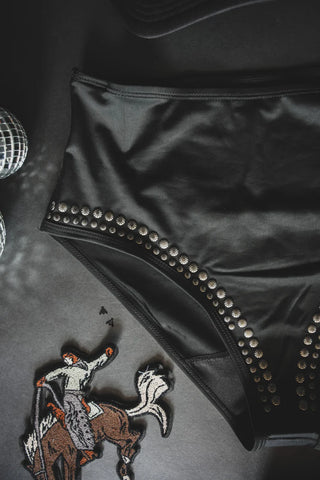 Here's a close-up of the CABO COWBOY BOTTOMS by 2 Fly, showcasing black swim bottoms adorned with silver studs along the sides. The backdrop features a glossy, reflective disco ball pattern and a fabric patch depicting a cowboy riding a bucking horse. This modest swimwear piece fuses edgy style with unique detailing.