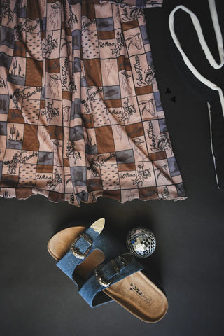 Flat lay of the Coastal Cowgirl Dress by 2 Fly, denim slide sandal, disco ball, and a black and white cactus object. The dress features western check print designs with cowboy themes like horses and cacti against a brown and pink checkered background.