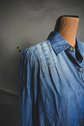 A close-up of the COWBOY BLUES chambray shirt by 2 Fly, showcasing its collared neckline and pleated shoulder design, displayed on a mannequin. The shirt features green and silver button details. The background is a plain, dark color.