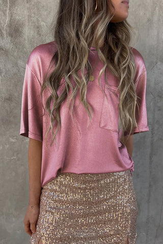 A person with long, wavy hair stands against a textured gray wall, wearing the Luca Satin Short Sleeve Pocket Blouse from Western Edge Boutique in a shiny pink hue, paired with a glittery gold skirt. Their outfit seamlessly combines breathable material for stylish comfort. The person's face is not visible.