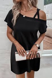 A woman in an off-shoulder ASYMMETRIC SHORT SLEEVE MINI DRESS from Western Edge Boutique holds a white clutch. She accessorizes with a necklace and watch, her long, wavy hair cascading over her shoulders. This fashion-forward look stands out against the light-colored wall with a glimpse of a couch in the background.