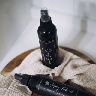 Two black room sprays from the Western Edge Boutique, labeled "Oh make your room smell oh sooo good... Room Spray" in the scents "Malagasy Teakwood" and "Sugared Pine," rest on a wooden tray with a textured beige cloth underneath. They are positioned on a white surface, suggesting a minimalist and cozy setting.