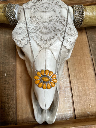 Mounted on a wooden wall, a decorated cow skull features an intricate lace pattern on its forehead and horns wrapped with metallic chains. Prominently displayed on the skull's forehead is the Amber Concho Pendant Necklace from Western Edge Boutique, showcasing a burnished silver flower design with a central eye.