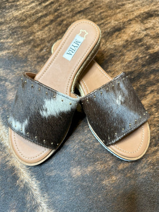 A pair of Taige Cowhide Leather Flats featuring fur-textured brown straps, accented with small golden studs along the edges. These durable flats have a beige sole and are branded with the name "Myra." The background showcases a similar fur texture that matches the flats.