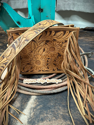 The Danni Tooled Leather Handbag by American Darling is displayed, showcasing its detailed, genuine leather adorned with intricate floral and leaf engravings. Perfectly sized, the bag features long fringe on the sides and an ornate strap. A "Western Edge Boutique" tag is placed in front, with a coiled rope partially visible beneath the bag.
