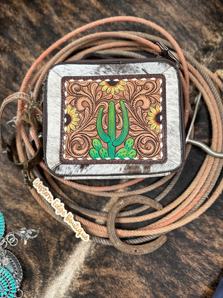 A square Carson Cowhide and Tooled Leather Travel Jewelry Case, crafted from genuine vegetable tanned cowhide and adorned with a detailed embossed design featuring a green cactus and sunflowers, is placed on a rustic background. This exquisite case from American Darling features durable zipper closures and is surrounded by coiled ropes and a turquoise earring, evoking a Western aesthetic. A tag reads "Western Lady Boutique.