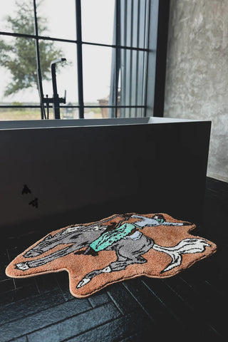 A modern black bathtub is situated on a glossy black tiled floor, in front of a large window. On the floor by the tub, there's a BRONC BUSTER rug by 2 Fly made of plush polyester, featuring a playful design of a rabbit wearing a green outfit. Natural light illuminates the room.