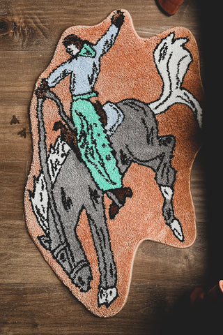 A colorful, irregularly shaped BRONC BUSTER rug from 2 Fly on a wooden floor. The plush polyester rug showcases a bold, cartoon-like illustration of a person in a green outfit riding a gray horse. The rider is depicted with a raised arm while the horse is in a dynamic pose. Dimensions available upon request.