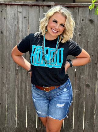 A smiling woman with short blonde hair stands in front of a wooden fence, wearing a "Howdy Black Turquoise Tee" from Western Edge Boutique, paired with denim shorts and striking turquoise jewelry. Her hands rest on her hips as she enjoys the outdoors.