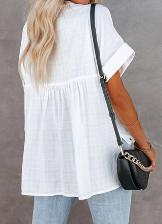 A woman with long blonde hair is wearing a loose white short-sleeved blouse, specifically the Aria Loose V Neck Lightweight Summer Women's Top by Pretty Bash, paired with light blue jeans and a black crossbody purse slung over her shoulder. The background is plain gray. This lightweight woven blouse features dolman sleeves and a gathered seam below the shoulders for a relaxed fit.