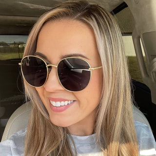 A woman with long blonde hair smiles in a car, wearing DAX Eyewear's stylish Georgie Gold Frame Sunglasses with UV protection. The round gold frames perfectly complement her look. Through the window, a fence and building are partially visible outside.
