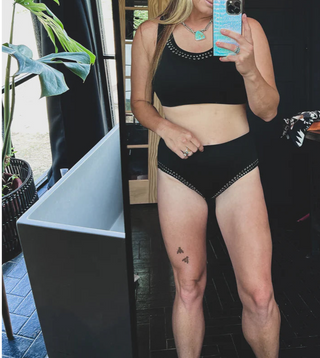 A person takes a mirror selfie in 2 Fly's CABO COWBOY BOTTOMS, a black, high-waisted bikini with decorative trim, pairing it confidently with silver studs. They have long hair and are wearing a turquoise necklace. Part of a plant and modern decor are visible in the background, along with a tattoo of three small butterflies on their thigh.