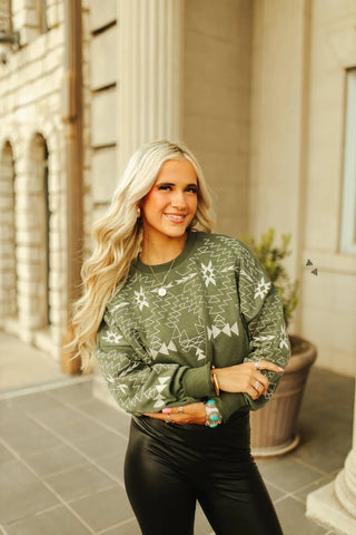 A woman with long blonde hair is wearing the Saddle Ranch Sweater by 2FLY, an olive green patterned sweater adorned with subtle embroidery. She pairs it with black leather pants and stands outside a stone building with arches and a large potted plant, smiling as she gazes into the distance.