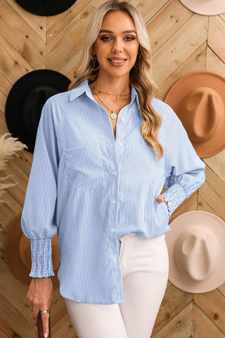 A woman with long blonde hair is smiling and wearing the Taylor Smocked Cuffed Striped Boyfriend Shirt with Pocket by Aapparella, paired with white pants. She stands in front of a wooden wall adorned with various hats in different colors.