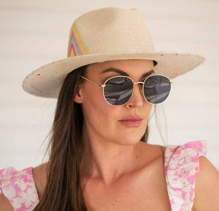 A woman with long brown hair wears a wide-brimmed straw hat and DAX Eyewear’s Georgie Sunglasses, featuring gold metal frames and UV-protective lenses. She's dressed in a pink and white floral top against a light, plain background.