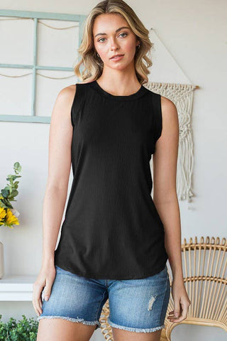 A woman with wavy blonde hair is wearing the Bella Black Ribbed Tank from Western Edge Boutique and blue denim shorts. She stands in a room with minimalistic decor, including light-colored walls, a wooden chair, and some plants. She gazes directly at the camera with a neutral expression.