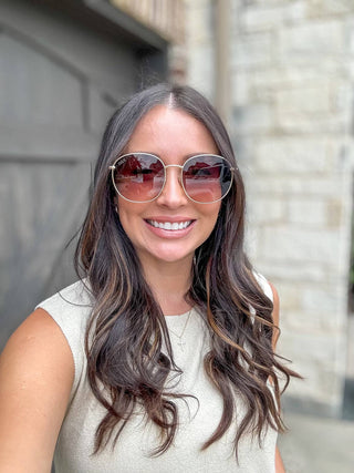 A woman with long, wavy brown hair and a sleeveless top smiles at the camera. She's wearing DAX Eyewear's Georgie sunglasses with gold frames offering UV protection. A stone wall and garage door are in the background.