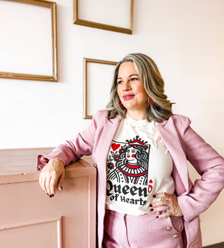 A person with shoulder-length gray hair leans on a dusty pink piece of furniture, wearing a pink suit and the "Queen of Hearts Graphic Tee" by Mugsby. Empty picture frames hang on the light wall behind, contrasting intriguingly with this rodeo queen's unique style.