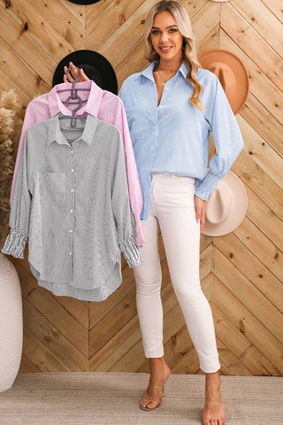 A woman with long blonde hair stands against a wooden chevron wall, wearing a Taylor Smocked Cuffed Striped Boyfriend Shirt with Pocket in light blue from Aapparella and white pants. She holds two additional Taylor Smocked Cuffed Striped Boyfriend Shirts, one gray and one pink, on hangers. Various hats are displayed on the wall.