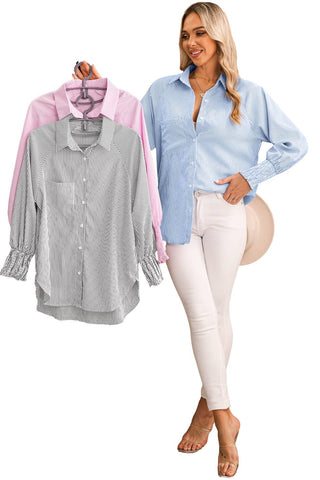 A woman with long blonde hair is standing and smiling, holding three striped shirts on hangers. She is wearing a light blue Taylor Smocked Cuffed Striped Boyfriend Shirt with Pocket by Aapparella, paired with white pants and beige heels. The shirts on the hangers are white with black stripes, pink with white stripes, and blue with white stripes.