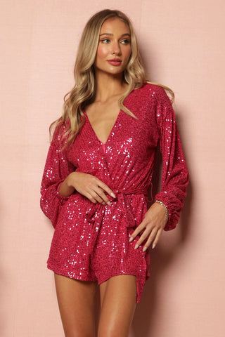 A woman with long blonde hair poses against a pink backdrop. She is wearing the Sheridan Sequin Romper from Blue Buttercup, a bright fuchsia sequin romper with long sleeves and a V-neck. Her hands rest casually at her waist, and she gazes slightly to the side, the shimmery sequins catching the light beautifully.