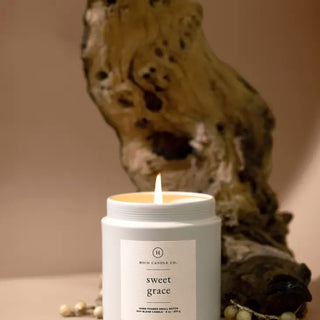 A lit Sweet Grace Candle 9 oz. from Hico Candle Co., housed in a white jar, is placed in front of a piece of driftwood. Made from soy wax, the candle emits a warm, soft glow with hints of passion fruit. The background has neutral tones, creating a serene and cozy atmosphere. Small decorative beads are arranged around the candle.